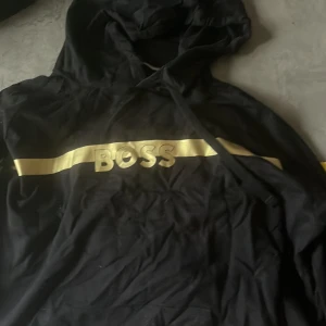 Svart hoodie från BOSS med gulddetalj - Svart hoodie från BOSS med en bred guldfärgad rand och BOSS-logga i guld över bröstet. Tröjan har huva och en klassisk, avslappnad passform. Perfekt för dig som gillar streetstyle och vill sticka ut med snygga detaljer. Inga skador 