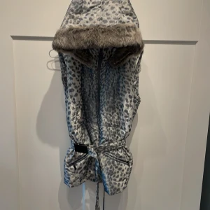 Leopardmönstrad dunväst med fuskpäls - Snygg dunväst i grått och svart leopardmönster med stor huva kantad av mjuk fuskpäls. Västen har dragkedja framtill, knytbälte i midjan och två fickor med dragkedja. Perfekt för att sticka ut och hålla värmen under kyliga dagar.