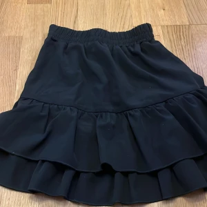 Svart volangkjol med resårmidja - Supersöt svart kortkjol med dubbla volanger och bred resår i midjan för en bekväm passform. Kjolen har en flowy känsla och passar perfekt till sneakers eller boots. Enkel att matcha och ger en cool vibe till din outfit.