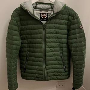 Grön Colmar pufferjacka med huva - Snygg grön pufferjacka från Colmar med huva och dragkedja framtill. Jackan har två fickor med dragkedja och diskreta logodetaljer på ärmen och nedtill. Perfekt för kyliga dagar och riktigt skön att bära. Fodrad insida i ljusgrått.