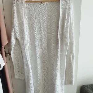 Vit lång kofta från H&M - Säljer en lång, öppen vit kofta från H&M med snyggt stickat mönster och långärmad design. Perfekt att slänga över en topp för en chill och stilren look. Koftan har ett diskret geometriskt mönster och är gjord i mjukt stickat material.