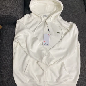 Vit hoodie från Lacoste XL - Snygg vit hoodie från Lacoste i regular fit med dragkedja framtill och klassisk krokodillogga på bröstet. Tröjan har huva med snörning och två fickor framtill. Fick den som present, men var tyvärr för stor. Storlek XL 