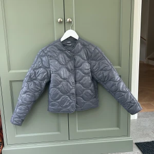 Grå/blå quiltad vår/höst jacka  - Quiltad grå jacka från Mango med coolt vågigt mönster. Jackan har rundad hals, tryckknappar framtill och lång ärm. Perfekt för lager-på-lager och ger en trendig vibe till din outfit. Materialet är syntet och jackan har en lätt pufferstil. !Aldrig använd!