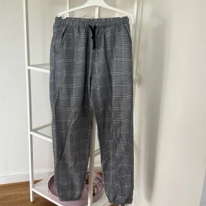 Grå rutiga mjukisbyxor från Shein - Grå mjukisbyxor från Shein med svartvitt rutigt mönster. Byxorna har elastisk midja med svart snörning och resår vid bensluten. Sköna och stilrena, perfekta för chill dagar. Tillverkade i mjukt material för extra komfort.