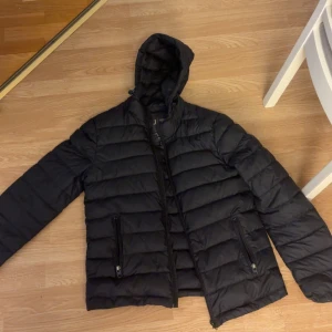 Svart dunjacka från Armani Jeans - Säljer en svart dunjacka från Armani Jeans med huva och dragkedja. Jackan har två fickor med dragkedja och är quiltad för extra värme. Perfekt för kalla dagar och har en clean, stilren look som funkar till det mesta.