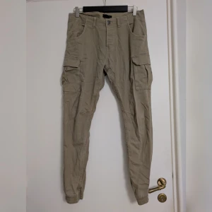 Beige cargobyxor - Snygga beige cargobyxor med flera fickor på sidorna och baktill. Byxorna har resår vid bensluten och är tillverkade i bomull med lite stretch för extra komfort. Perfekta för en avslappnad och trendig look. De är bara skrynkliga, men i bra skick. Insidan av fickorna är noppiga, men det påverkar inte utseendet. 