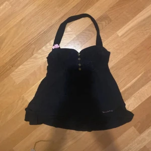 Svart babydolltopp med knappar - Supersöt svart babydolltopp som är hemmasydd men bondelid med breda axelband och dekorativa knappar framtill. Toppen har en hjärtformad urringning och en liten rosa blomdetalj på axelbandet. Perfekt för att skapa en cool och enkel look.