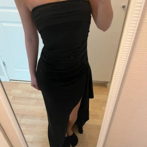 Svart axelbandslös långklänning - Säljer en svart axelbandslös långklänning med tight passform och snygg slits framtill. Klänningen har draperade detaljer över bysten och midjan som ger en elegant look. Perfekt för dig som vill sticka ut på festen!