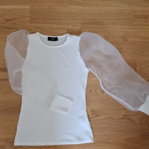 Vit ribbad topp med puffärmar Sisters Point - Superfin vit ribbad topp från Sisters Point med transparenta puffärmar i mesh. Toppen har rund halsringning och långa ärmar med breda muddar. Perfekt för dig som vill sticka ut med en trendig och elegant look.