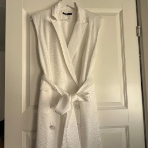 Vit omlottklänning från Gina Tricot - Snygg vit omlottklänning från Gina Tricot med markerad krage, knytband i midjan och stora klädda knappar framtill. Klänningen är ärmlös och har en lätt glansig yta som ger en lyxig känsla. Perfekt för dig som vill sticka ut med en stilren look.