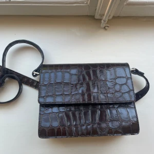 Mörkbrun axelväska från Ganni - Säljer en snygg mörkbrun axelväska från Ganni i glansigt skinn med croco-mönster. Väskan har en stilren, rektangulär form och justerbar axelrem. Perfekt för dig som vill ha en trendig och tidlös accessoar med lite edge.