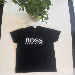 Svart Hugo Boss t-shirt med logga - Svart t-shirt från Hugo Boss med stor vit BOSS-logga framtill. Klassisk rund hals och korta ärmar. Skön passform och tillverkad i mjuk bomull, perfekt för en clean och stilren look. Den saknar dock neck tagen.