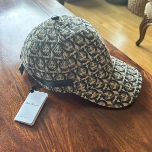 Mönstrad keps från Salvatore Ferragamo - Snygg keps från Salvatore Ferragamo med svart och beige mönster över hela kepsen. Klassisk böjd skärm och justerbar rem bak. Tillverkad i Italien och i bomullsmaterial. Perfekt för dig som vill sticka ut med en exklusiv accessoar.