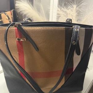 Rutig axelväska från B U r B E R R Y - Snygg axelväska  med klassiskt rutigt mönster i beige, svart, rött och brunt. Väskan har svarta skinnhandtag, guldfärgade metalldetaljer och dragkedja upptill. Insidan är rymlig med flera fack för smart organisering.