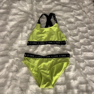 Grön bikini - Sportig grön bikini från Ü RüKie med svarta elastiska band med logga. Toppen har korsade axelband och hög hals, medan trosan har en klassisk passform. Materialet är glansigt och stretchigt, perfekt för stranden eller poolen. Den har en lite skrikigare färg i verkligenheten, tog bilderna på kvällen så skicka privat ifall ni vill ha bättre bilder! Storlek 134/140 men passar definitivt större. Inga fläckar över huvud taget!