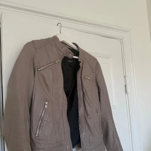 Beige skinnjacka Rockandblue, stl 38 - Snygg beige skinnjacka från Rockandblue med coola detaljer som dragkedjor på bröstet och fickorna samt ståkrage med tryckknapp. Jackan har en klassisk bikerstil och är perfekt för dig som vill ha en stilren look med edge.