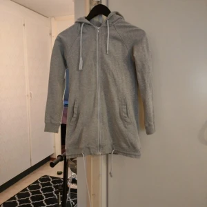 Lång grå hoodie med dragkedja - Lång och mysig grå hoodie med huva och snörning. Tröjan har hel dragkedja framtill, två fickor och ribbade muddar vid ärmslut. Perfekt för chill dagar eller när du vill ha något bekvämt och stilrent.