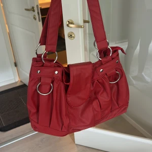 Röd axelväska med stora ringdetaljer - Säljer en snygg röd axelväska med breda axelband och stora silvriga ringdetaljer. Väskan har veckad design och stängs med ett lock framtill. Perfekt storlek för att få plats med allt du behöver och ger en cool statement-look.