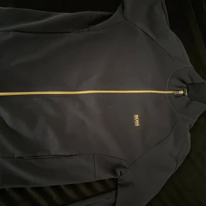 Svart zip-jacka från BOSS - Svart jacka från BOSS med guldfärgad dragkedja och logga på bröstet. Jackan har hög krage, långa ärmar och är tillverkad i ett mjukt material som känns skönt mot huden. 