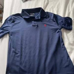 Marinblå pikétröja från Ralph Lauren - Snygg marinblå pikétröja från Ralph Lauren med klassisk krage och korta ärmar. Tröjan har den ikoniska röda polospelaren broderad på bröstet och är tillverkad i mjuk bomull. Perfekt för en clean och stilren look.