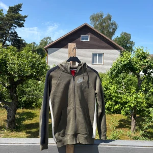Olivgrön Nike hoodie med dragkedja XS - Nike Air hoodie i olivgrönt med svarta och vita detaljer längs ärmarna. Tröjan har huva, hel dragkedja och två fickor framtill. Perfekt för chill dagar eller till träning. Klassisk Nike-logga på bröstet och mjukt material. Passar S bra 