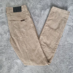 Beiga manchesterbyxor från Nudie Jeans - Snygga beiga manchesterbyxor från Nudie Jeans med klassisk femficksdesign och raka ben. Byxorna har diskret logga på bakfickan och är tillverkade i mjuk manchester som ger en chill och stilren vibe. OBS! En knap har ramlat av