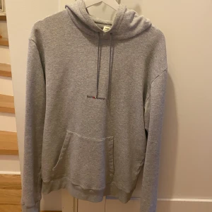 Grå hoodie från Saint Laurent - Säljer en stilren grå hoodie från Saint Laurent Paris. Tröjan har huva med dragsko, magficka och diskret logga på bröstet. Perfekt för en avslappnad och trendig look. Inge kvitto finns därför säljs billigt.