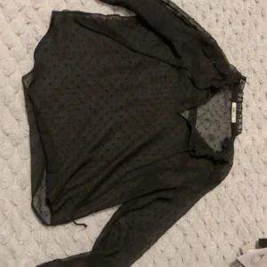 Svart transparent blus från Gina Tricot - Säljer en svart transparent blus från Gina Tricot i storlek 36. Blusen har lång ärm och ett diskret prickigt mönster i tyget. Perfekt att styla lager på lager för en trendig look.