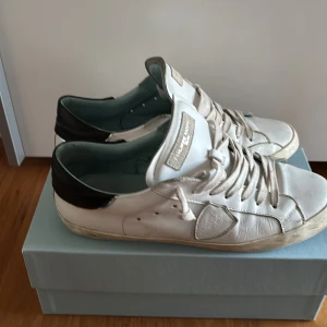 Vita sneakers från Philippe Model  - Säljer ett par vita sneakers från Philippe Model med svarta detaljer på hälen och bruna gummisulor. Skorna har snörning och en klassisk låg profil. Perfekta för en stilren och avslappnad look.