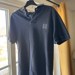 Mörkblå pikétröja från Stone Island - Säljer en mörkblå pikétröja från Stone Island med klassisk krage och korta ärmar. Tröjan har en broderad logotyp på bröstet och knappar vid halsen. Perfekt för en stilren och avslappnad look.