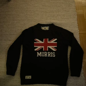 Svart stickad tröja med Union Jack från Morris - Svart stickad tröja från Morris med stor Union Jack-flagga i rött, vitt och blått på bröstet samt texten 'MORRIS' under. Rund halsringning och långa ärmar. Perfekt för dig som gillar klassisk brittisk stil. Perfekt till våren och sena sommarkvällar.