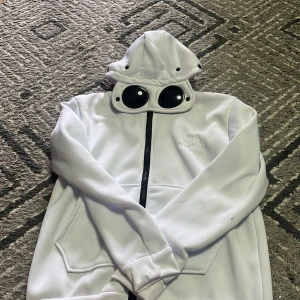 Vit hoodie med glasögon i huvan från C.P. Company - Säljer en vit hoodie från C.P. Company med unika glasögon i huvan och dragkedja framtill. Tröjan har broderad logga på bröstet och ribbade muddar vid ärmslut och nederkant. Perfekt för dig som vill sticka ut!