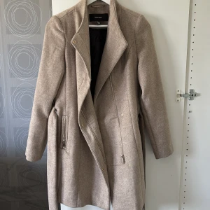 Beige kappa från Vero Moda - Stilren beige kappa från Vero Moda med bälte i midjan och långa ärmar.
