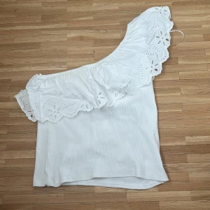 Vit oneshoulder topp med volang från ZARA - Säljer en vit oneshoulder topp från ZARA med bred volangkant och broderade blommor. Toppen är ärmlös och har en snygg asymmetrisk design som ger en trendig look.