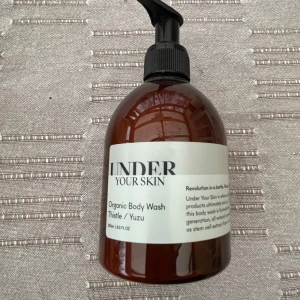Oöppnad Under Your Skin Organic Body Wash  - En brun plastflaska med pump, innehållande en ekologisk body wash från Under Your Skin med doft av tistel och yuzu. Flaskan har en vit etikett med svart text och rymmer 250 ml.