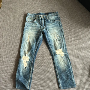 Ljusblå slitna jeans från Tiger of Sweden - Säljer ett par ljusblå jeans från Tiger of Sweden med rejält slitna detaljer och hål på båda benen. Klassisk femficksmodell med raka ben och snygg tvätt. Perfekta för en avslappnad och trendig look.