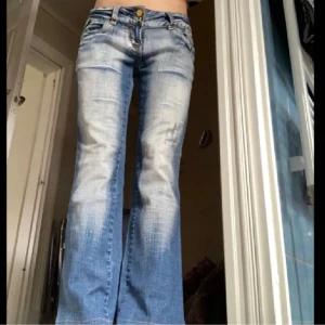 Blå bootcut jeans från Reals - Säljer ett par blå bootcut jeans från Reals med slitna detaljer och ljus tvätt. Jeansen har klassiska fem fickor, låg midja och gylf med knapp. Märkespatch bak i midjan och snygga kontrastsömmar.