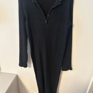 Svart ribbad långärmad klänning - Svart ribbad långärmad klänning med krage och knappar framtill. Klänningen har en kroppsnära passform och är tillverkad i ett stretchigt material som ger en snygg siluett. Perfekt för dig som gillar en stilren och enkel look. Har en super liten fläck på kjolen men går bort bara utav att riva med fingret! 