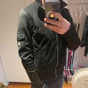 Svart bomberjacka med detaljer - Säljer en svart bomberjacka med dragkedja och fickor på ärmen. Jackan har ribbade muddar och en modern, normal passform. Ärmarna har en snygg textur som ger extra stilpoäng. Perfekt för dig som vill ha en stilren och trendig jacka.