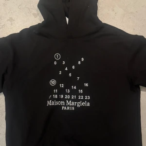 Svart hoodie från Maison Margiela - Säljer en svart hoodie från Maison Margiela med det ikoniska numermotivet och loggan tryckt på bröstet. Tröjan har långa ärmar och klassisk huva. Perfekt för dig som gillar stilrena designer med en twist. Använd två gånger för den var lite för stor