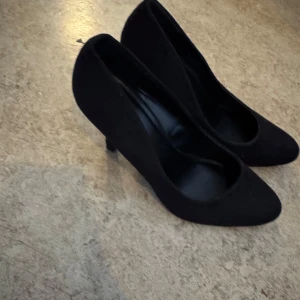 Svarta pumps med klack - Säljer ett par klassiska svarta pumps med rund tå och hög klack. Skorna har en stilren design och är tillverkade i ett material som liknar mocka. Perfekta för dig som vill ha en tidlös och elegant sko, perfekt för balen.