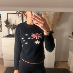 Mörkblå stickad tröja med australiensisk flagga - Mörkblå stickad tröja med rund hals och lång ärm. Framtill finns ett stort motiv av Australiens flagga i rött, vitt och blått. Tröjan är croppad och har ribbade muddar vid ärmslut och hals. Tröjan är ifrån Holebrook Sweden 