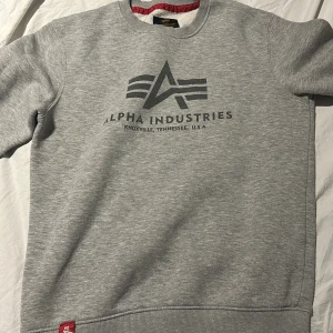 Grå sweatshirt från Alpha Industries - Säljer en grå sweatshirt från Alpha Industries med tryckt logga och text på bröstet. Tröjan har rund halsringning, ribbade muddar och mjuk insida. Perfekt för en avslappnad stil.