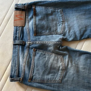 Blå jeans från Nudie Jeans - Säljer ett par klassiska blå jeans från Nudie Jeans med snygga slitningar och kontrastsömmar. Jeansen har fem fickor, normal passform med en super låg midja och raka ben. Perfekta för en stockholms stil.