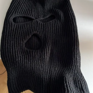 Svart stickad balaclava - Svart ribbstickad balaclava med tre hål för ögon och mun. Perfekt för kalla dagar eller utomhusaktiviteter. Materialet är mjukt och stretchigt för en bekväm passform.