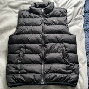 Svart dunväst med dragkedja - Snygg svart dunväst med hög krage och dragkedja framtill. Västen har två snedställda fickor på sidorna och quiltad design med breda kanaler. Perfekt för lager-på-lager under kyliga dagar.