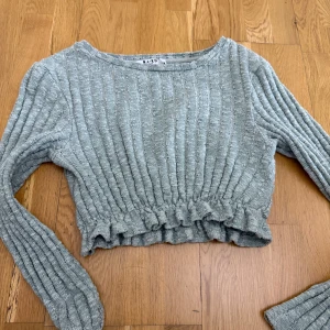 Ljusgrå ribbad croppad tröja från NA-KD - Superfin ljusgrå ribbad tröja från NA-KD med croppad passform och volangkant nedtill. Tröjan har långa ärmar och är perfekt för lager-på-lager. Snygg och enkel att matcha till olika outfits.