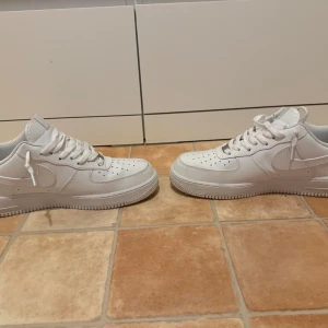 Vita Nike Air Force 1 sneakers - Klassiska helvita Nike Air Force 1 sneakers med lågt skaft och vit sula. Skorna har snörning och är tillverkade i skinn med perforerade detaljer på tån för extra ventilation. Perfekta för en clean och stilren look.