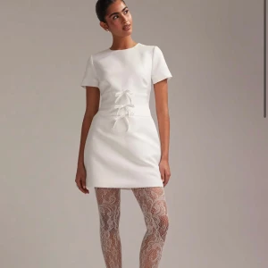 Vit kortklänning med rosetter och öppen rygg - Stilren vit kortklänning med korta ärmar, tre dekorativa rosetter framtill och en snygg öppen rygg. Klänningen har en rak passform och är tillverkad i ett slätt material som ger en elegant känsla. Säljer på grund av missad returdag, nypris är 1800kr 