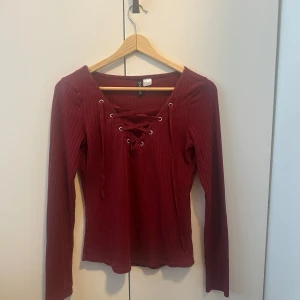 Vinröd ribbad topp med snörning - Vinröd långärmad topp från H&M med ribbad struktur och snygg snörning framtill vid halsen. Toppen har en v-ringad design och är figurnära i passformen. Perfekt för att lyfta din outfit med en trendig detalj.